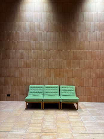 A row of three green upholstered chairs with button details sits against a brown tiled wall. The floor is also tiled, matching the wall color. The lighting casts a gentle shadow above the chairs, giving the setting a peaceful and minimalist feel.
