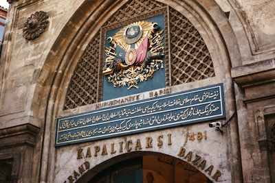 An ornate archway features a richly detailed emblem set against a blue background, depicting symbols and an intricate design with gold, red, and metallic elements. Below, a sign with elegant script is mounted on stone, surrounded by a patterned stone texture. The entrance is labeled as 'Kapalıçarşı' and 'Grand Bazaar' with the date 1461.