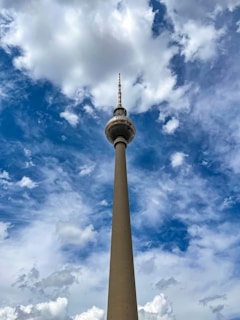 A tall communications tower rises against a vibrant sky filled with fluffy, white clouds. The structure features a spherical section near the top with an antenna extending upward, set against a backdrop of deep blue contrasted by the bright clouds.