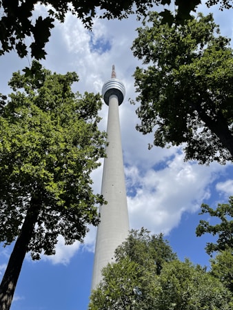 A tall communication tower rises into the sky, surrounded by lush green trees. The angle from below highlights the structure's height and sleek design. Fluffy white clouds and patches of blue sky provide a natural backdrop.