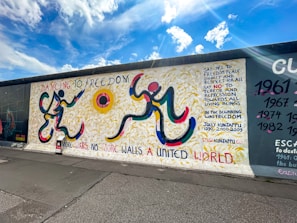 A vibrantly painted section of a wall features abstract human figures rendered with bold, curved lines in yellow, red, blue, and black. The background showcases yellow strokes resembling wheat fields. Text in various colors includes phrases advocating for freedom, peace, dignity, and unity. The sky is bright with fluffy clouds, adding to the sense of openness.