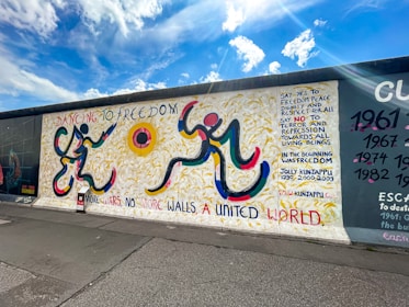 A vibrantly painted section of a wall features abstract human figures rendered with bold, curved lines in yellow, red, blue, and black. The background showcases yellow strokes resembling wheat fields. Text in various colors includes phrases advocating for freedom, peace, dignity, and unity. The sky is bright with fluffy clouds, adding to the sense of openness.