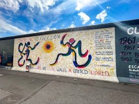 A vibrantly painted section of a wall features abstract human figures rendered with bold, curved lines in yellow, red, blue, and black. The background showcases yellow strokes resembling wheat fields. Text in various colors includes phrases advocating for freedom, peace, dignity, and unity. The sky is bright with fluffy clouds, adding to the sense of openness.
