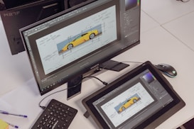 A desktop setup featuring two monitors displaying a design software with a digital sketch of a sports car. The primary monitor is elevated on a stand, while the secondary is a drawing tablet laying on the desk. A wireless keyboard, mouse, and pens are positioned nearby.