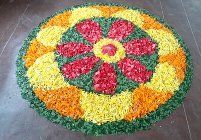 A vibrant circular floral arrangement made with petals of various colors including red, yellow, orange, and green. The pattern forms a symmetrical design with a central red flower surrounded by alternating sections of yellow and orange petals. It is placed on a brown tiled floor.