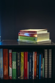 A collection of self-help and motivational books are neatly organized on a black bookshelf. On the top shelf, there is a small stack of books including 'Tuesdays with Morrie', 'The Magic of Thinking Big', 'Think and Grow Rich', and 'The Power of Now'. The bottom shelf displays a variety of books such as 'The Alchemist', 'Freedom from the Known', and 'The 7 Habits of Highly Effective People'. The setup suggests a personal library focused on personal development.