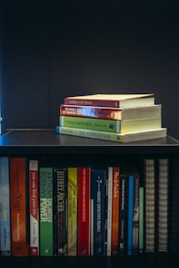 A collection of self-help and motivational books are neatly organized on a black bookshelf. On the top shelf, there is a small stack of books including 'Tuesdays with Morrie', 'The Magic of Thinking Big', 'Think and Grow Rich', and 'The Power of Now'. The bottom shelf displays a variety of books such as 'The Alchemist', 'Freedom from the Known', and 'The 7 Habits of Highly Effective People'. The setup suggests a personal library focused on personal development.