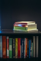 A collection of self-help and motivational books are neatly organized on a black bookshelf. On the top shelf, there is a small stack of books including 'Tuesdays with Morrie', 'The Magic of Thinking Big', 'Think and Grow Rich', and 'The Power of Now'. The bottom shelf displays a variety of books such as 'The Alchemist', 'Freedom from the Known', and 'The 7 Habits of Highly Effective People'. The setup suggests a personal library focused on personal development.
