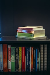A collection of self-help and motivational books are neatly organized on a black bookshelf. On the top shelf, there is a small stack of books including 'Tuesdays with Morrie', 'The Magic of Thinking Big', 'Think and Grow Rich', and 'The Power of Now'. The bottom shelf displays a variety of books such as 'The Alchemist', 'Freedom from the Known', and 'The 7 Habits of Highly Effective People'. The setup suggests a personal library focused on personal development.