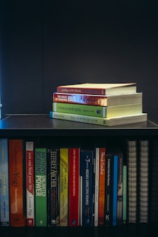 A collection of self-help and motivational books are neatly organized on a black bookshelf. On the top shelf, there is a small stack of books including 'Tuesdays with Morrie', 'The Magic of Thinking Big', 'Think and Grow Rich', and 'The Power of Now'. The bottom shelf displays a variety of books such as 'The Alchemist', 'Freedom from the Known', and 'The 7 Habits of Highly Effective People'. The setup suggests a personal library focused on personal development.