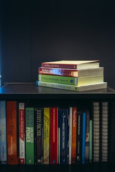 A collection of self-help and motivational books are neatly organized on a black bookshelf. On the top shelf, there is a small stack of books including 'Tuesdays with Morrie', 'The Magic of Thinking Big', 'Think and Grow Rich', and 'The Power of Now'. The bottom shelf displays a variety of books such as 'The Alchemist', 'Freedom from the Known', and 'The 7 Habits of Highly Effective People'. The setup suggests a personal library focused on personal development.