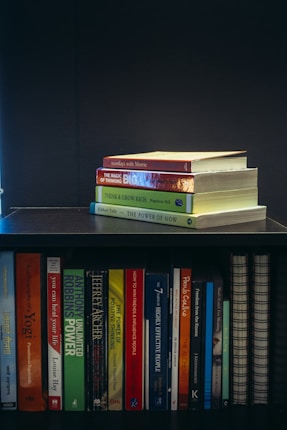 A collection of self-help and motivational books are neatly organized on a black bookshelf. On the top shelf, there is a small stack of books including 'Tuesdays with Morrie', 'The Magic of Thinking Big', 'Think and Grow Rich', and 'The Power of Now'. The bottom shelf displays a variety of books such as 'The Alchemist', 'Freedom from the Known', and 'The 7 Habits of Highly Effective People'. The setup suggests a personal library focused on personal development.