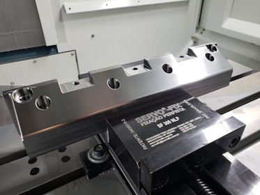 A metal workpiece with a precision-machined surface is clamped in a vise. The workpiece features several circular holes and notches. The vise is metallic with engraved text and is mounted on a flat surface inside a machine shop or manufacturing environment.
