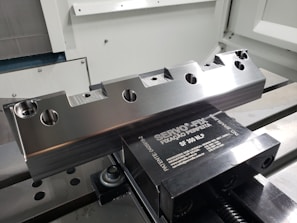 A metal workpiece with a precision-machined surface is clamped in a vise. The workpiece features several circular holes and notches. The vise is metallic with engraved text and is mounted on a flat surface inside a machine shop or manufacturing environment.