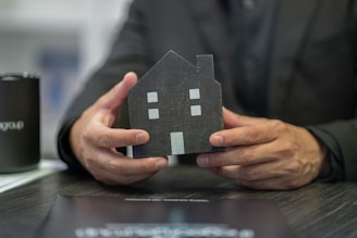 A person dressed in dark clothing is holding a small, black, house-shaped model with white windows and a door. The scene includes a black mug with text and some documents or papers on a dark wooden table.