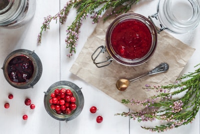 A jar of cranberry jam is placed on a piece of brown paper alongside a decorative spoon. Surrounding the jar are small bowls, one filled with whole cranberries and the other with more jam. Sprigs of green foliage and small pink flowers are artfully arranged around the jars, all set on a white wooden surface.