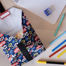 A colorful pouch decorated with various words and symbols is placed next to a lined notebook. Several colored pencils, including a teal pen and a small blue sharpener, are arranged nearby. The pouch appears to contain a snack, perhaps a cookie or a sandwich.