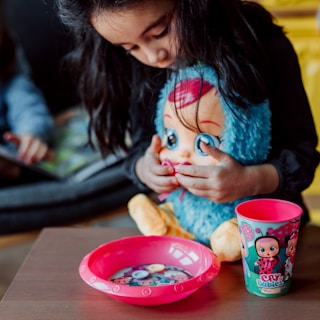 A young child holds a plush doll with oversized eyes and colorful hair, accompanied by a bright pink bowl and cup on a wooden surface. The cup features cartoon characters, adding a playful theme.
