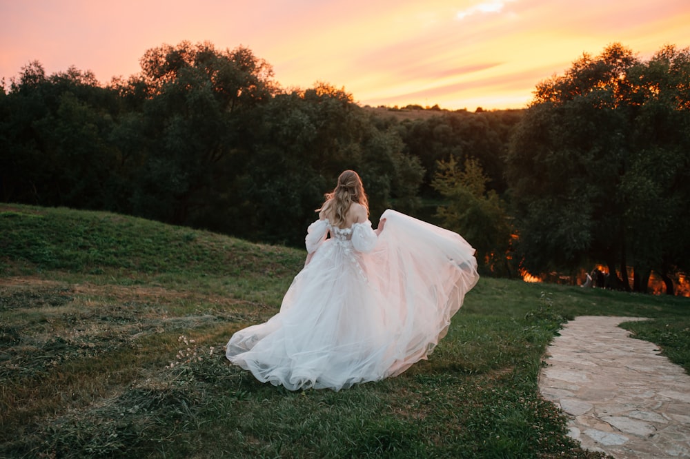 a person in a white dress sitting on a grassy hill with trees and a sunset