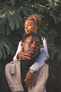 A young woman stands behind a young man, gently wrapping her arm around his shoulders. Both are dressed stylishly, with the woman sporting sunglasses on her head and glitter makeup, while the man wears a beige sleeveless jacket and a black turtleneck. They are surrounded by large green tropical leaves, creating a natural backdrop.