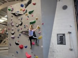 A person is climbing an indoor bouldering wall covered with multicolored climbing holds. The facility has a variety of holds in different shapes and sizes, attached to a light gray wall. The climber is mid-action, wearing athletic clothing. A chalkboard displays different grades, presumably indicating difficulty levels, and a clock is mounted on the wall.