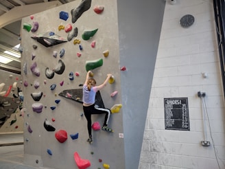 A person is climbing an indoor bouldering wall covered with multicolored climbing holds. The facility has a variety of holds in different shapes and sizes, attached to a light gray wall. The climber is mid-action, wearing athletic clothing. A chalkboard displays different grades, presumably indicating difficulty levels, and a clock is mounted on the wall.