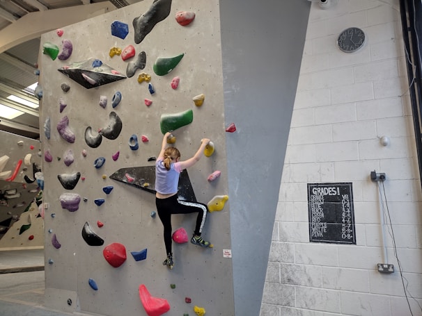 A person is climbing an indoor bouldering wall covered with multicolored climbing holds. The facility has a variety of holds in different shapes and sizes, attached to a light gray wall. The climber is mid-action, wearing athletic clothing. A chalkboard displays different grades, presumably indicating difficulty levels, and a clock is mounted on the wall.