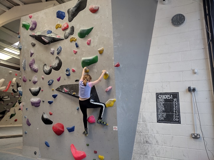 A person is climbing an indoor bouldering wall covered with multicolored climbing holds. The facility has a variety of holds in different shapes and sizes, attached to a light gray wall. The climber is mid-action, wearing athletic clothing. A chalkboard displays different grades, presumably indicating difficulty levels, and a clock is mounted on the wall.