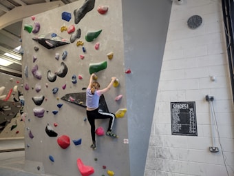 A person is climbing an indoor bouldering wall covered with multicolored climbing holds. The facility has a variety of holds in different shapes and sizes, attached to a light gray wall. The climber is mid-action, wearing athletic clothing. A chalkboard displays different grades, presumably indicating difficulty levels, and a clock is mounted on the wall.