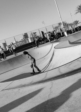 A black and white photograph captures a skater in motion within a skate park. The skater is wearing jeans and a shirt, navigating the curves of an empty concrete bowl. In the background, a crowd of people stands and sits near bicycles along a fence, with city buildings partially visible beyond. Palm trees and light poles add to the urban setting.