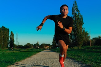 A person in athletic wear is sprinting down a gravel path surrounded by greenery. Tall trees and utility poles are visible in the background under a vibrant blue sky. The individual is focused, exhibiting determination.