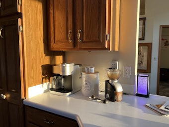A kitchen counter with wooden cabinets above. On the counter, there is a coffee machine, a ceramic canister, a coffee grinder, a smartphone on a stand, and a few small objects including two spoons. Sunlight is coming in, casting shadows, creating a warm and cozy ambiance.