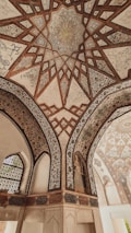 Intricate geometric patterns adorn the ceiling, showcasing detailed artwork with a combination of star-like shapes and floral motifs. The arches feature ornate designs, with Arabic calligraphy and a painted figure on the wall. The overall architectural style is historical and elegant.