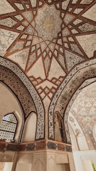 Intricate geometric patterns adorn the ceiling, showcasing detailed artwork with a combination of star-like shapes and floral motifs. The arches feature ornate designs, with Arabic calligraphy and a painted figure on the wall. The overall architectural style is historical and elegant.