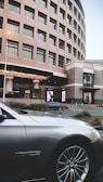 A modern, curved building with multiple windows stands in the background while a luxury car is parked in the foreground. Large digital advertisements are displayed on the building, and there are some bushes and parking cones around the area.