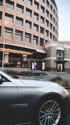 A modern, curved building with multiple windows stands in the background while a luxury car is parked in the foreground. Large digital advertisements are displayed on the building, and there are some bushes and parking cones around the area.