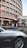 A modern, curved building with multiple windows stands in the background while a luxury car is parked in the foreground. Large digital advertisements are displayed on the building, and there are some bushes and parking cones around the area.