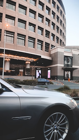 A modern, curved building with multiple windows stands in the background while a luxury car is parked in the foreground. Large digital advertisements are displayed on the building, and there are some bushes and parking cones around the area.