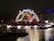 A brightly lit circus tent is set up on the water's edge at night, adorned with strings of decorative lights. The tent features a pointed top with red and yellow colors and a prominent sign saying 'CONELLI'. The reflection of the lights shimmers on the water surface, creating a festive atmosphere.