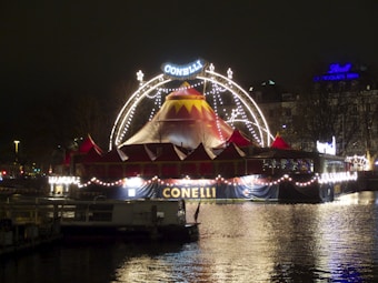A brightly lit circus tent is set up on the water's edge at night, adorned with strings of decorative lights. The tent features a pointed top with red and yellow colors and a prominent sign saying 'CONELLI'. The reflection of the lights shimmers on the water surface, creating a festive atmosphere.