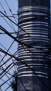 A metal pole wrapped with several layers of silver tape is surrounded by a complex network of black wires and cables extending in various directions against a light blue sky. The lower part of the pole shows some foliage in the background.
