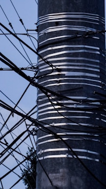 A metal pole wrapped with several layers of silver tape is surrounded by a complex network of black wires and cables extending in various directions against a light blue sky. The lower part of the pole shows some foliage in the background.