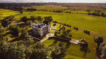 A large, elegant mansion set amidst expansive, lush green lawns and fields. The building is surrounded by trees and small garden areas, with a driveway leading up to it. Light and shadows create a serene atmosphere in the landscape, suggesting a peaceful rural setting.