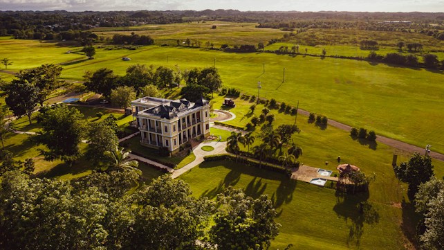 A large, elegant mansion set amidst expansive, lush green lawns and fields. The building is surrounded by trees and small garden areas, with a driveway leading up to it. Light and shadows create a serene atmosphere in the landscape, suggesting a peaceful rural setting.