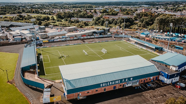 An aerial view of a football stadium, showing a well-maintained green pitch with goalposts. The stands are empty, and there are a few people visible on the field. Surrounding the stadium are various buildings, roads, and a parking area with several buses parked. In the background, there are trees and residential areas, and the sky is clear.