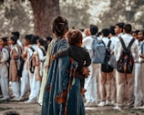A woman holding a child stands with her back to the camera, watching a group of students in school uniforms. The students are gathered in a loose formation, wearing light-colored uniforms with backpacks. The woman is wrapped in a colorful shawl with floral patterns, and the scene takes place in a park-like setting with trees in the background.