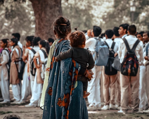 A woman holding a child stands with her back to the camera, watching a group of students in school uniforms. The students are gathered in a loose formation, wearing light-colored uniforms with backpacks. The woman is wrapped in a colorful shawl with floral patterns, and the scene takes place in a park-like setting with trees in the background.