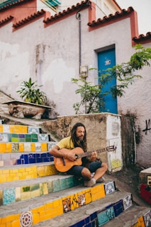 A man with dreadlocks is sitting on colorful tiled stairs playing an acoustic guitar. The stairs feature vibrant patterns in yellow, blue, and green. A blue door and green plants are in the background, with weathered walls surrounding the area.