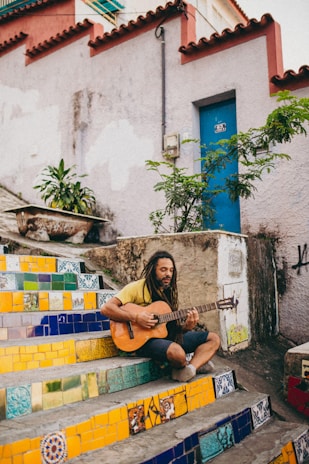 A man with dreadlocks is sitting on colorful tiled stairs playing an acoustic guitar. The stairs feature vibrant patterns in yellow, blue, and green. A blue door and green plants are in the background, with weathered walls surrounding the area.