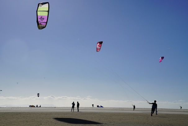 Several people are engaged in kiteboarding on a wide, sandy beach. The sky is clear and blue, with a few colorful kites flying high. The beach is open and expansive, with a calm and relaxed atmosphere.
